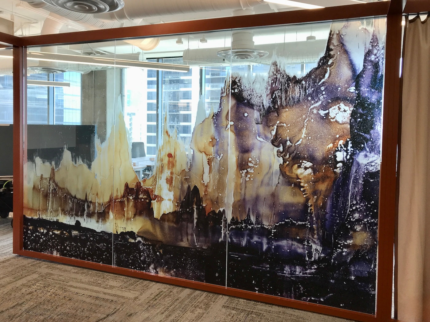 2. Large glass partition framed in rust-orange displaying Mystic Mountains by Jude Chase, a dynamic mountain-inspired composition in deep browns, golds, and purples. Transparent sections allow partial views of the office beyond, creating a layered, atmospheric effect.
