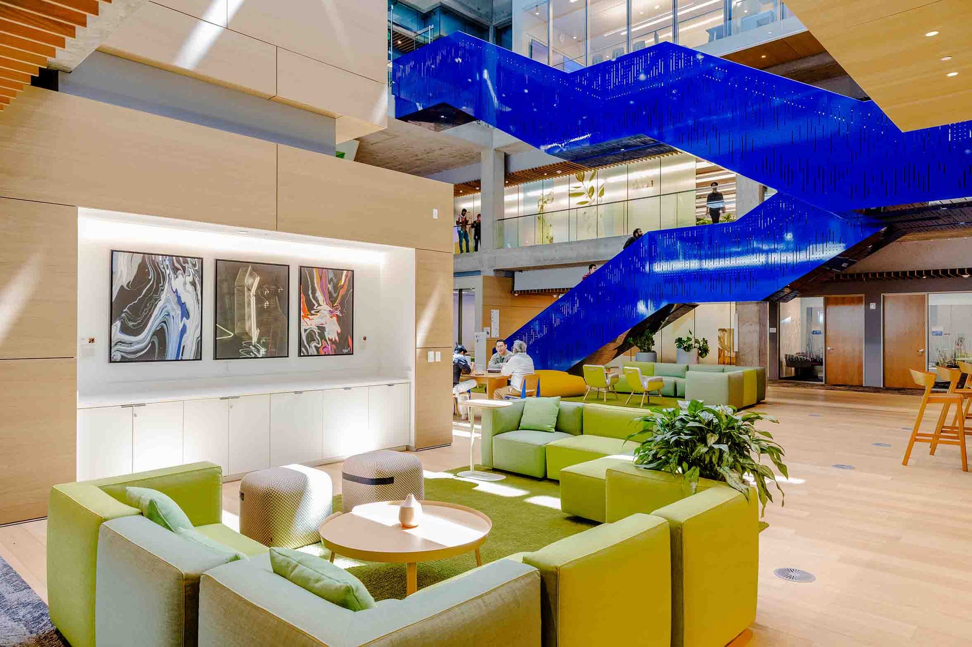 Three print reproductions hang in an open space on the second floor of MTV 22. From left to right: Floral Kaleidoscope by Sylvia Burnstein, Jewel Damiano Austin, Neon Lights by Sylvia Burnstein. The artworks’ dark backgrounds and fluid neon colors visually ground the open area, which features natural light, lime green furniture, plywood walls and floors, and a cobalt blue staircase. 