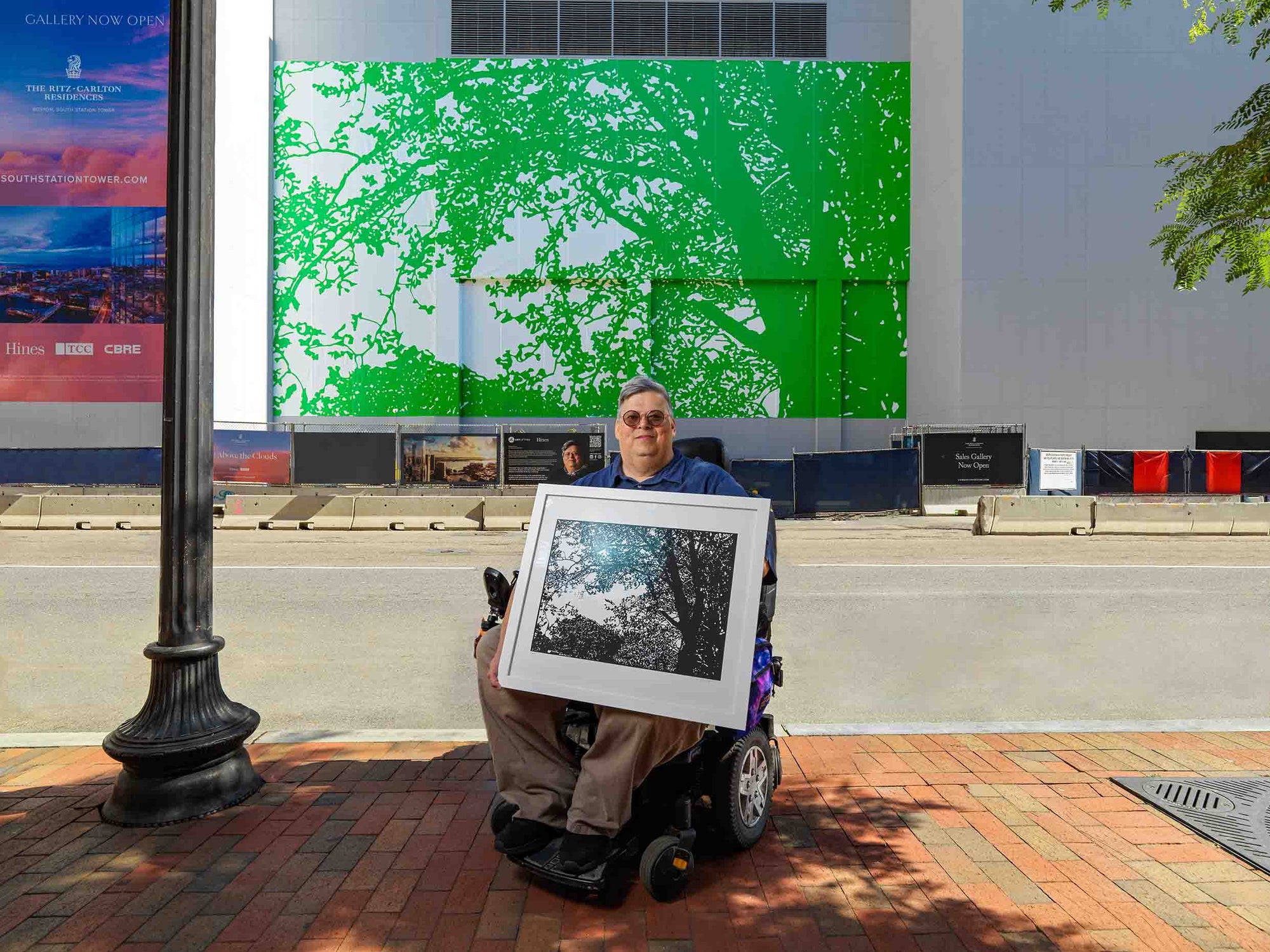 Person holding a framed picture outdoors with a large green and white artwork on a building wall.