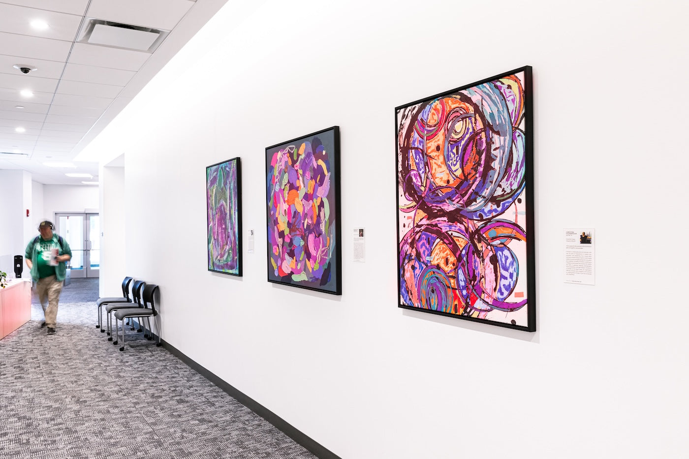 Vibrant, abstract prints line a white corridor. Colorful Chaotic World 3 (right) by Christina Culverhouse shows layered purple shapes, the middle work, Feelings: Uncategorized, by Madison Elyse Rubenstein combines overlapping patches of pink, orange, and teal. 