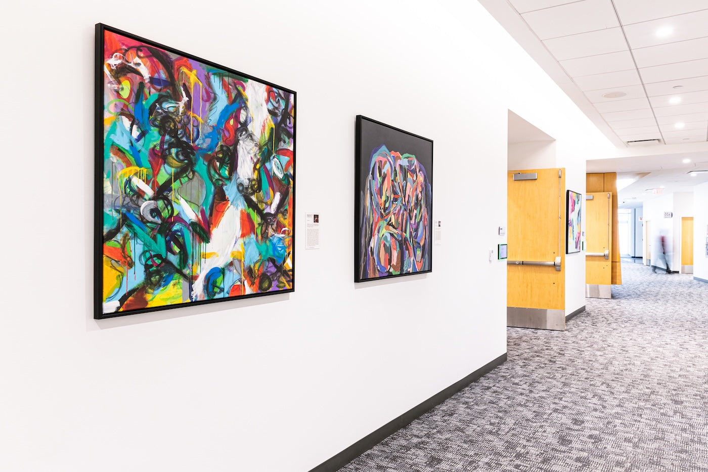 Two large abstract paintings hang in a bright hallway. Always In (left) by Madison Elyse Rubenstein bursts with swirling strokes of red, turquoise, black, and yellow, while Relentless Spirit of Positive Transformation (right) by Quána Madison features layered circular forms in pink, purple, and blue on a dark background.