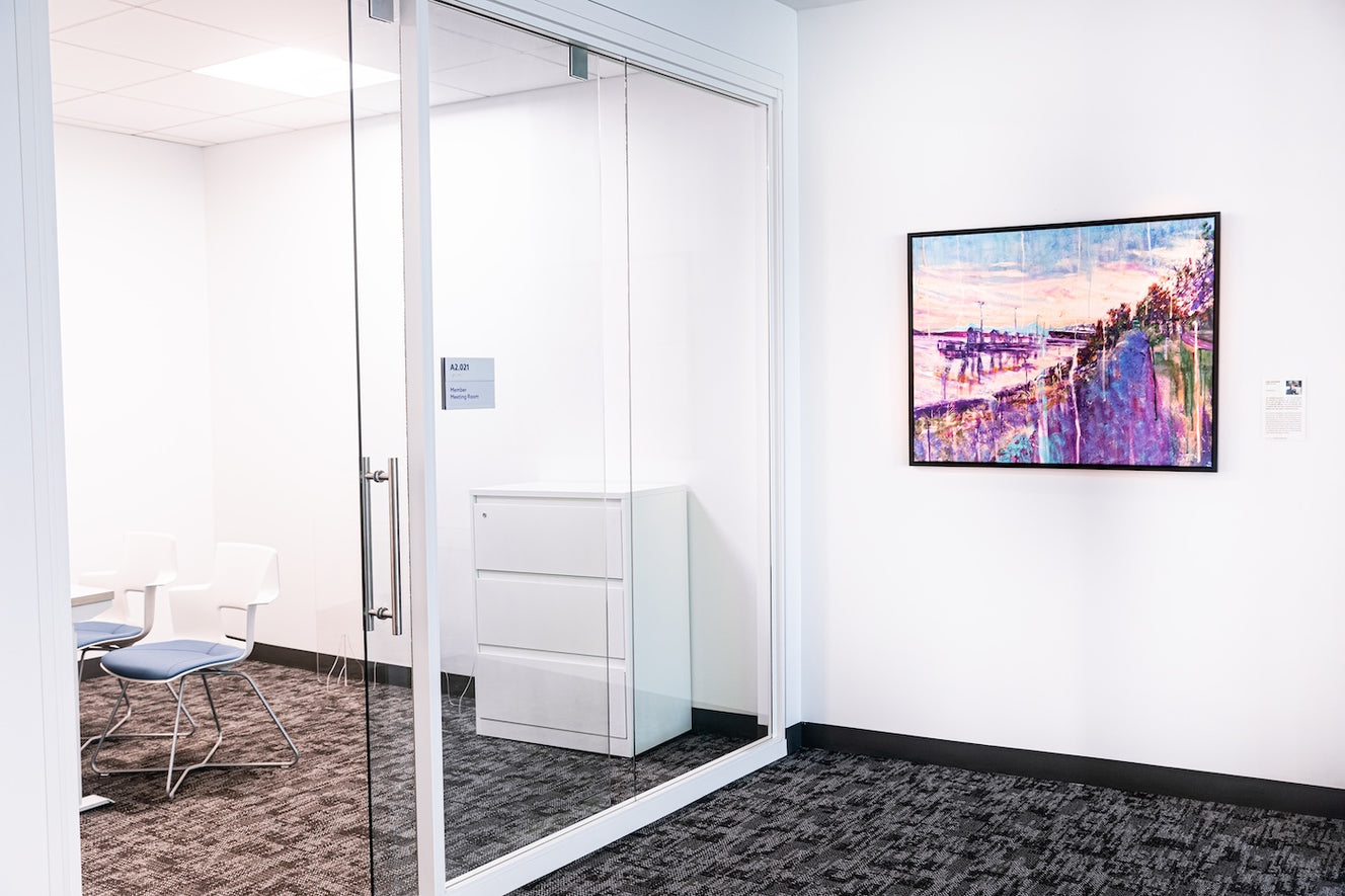  Displayed beside a glass-walled meeting room, Incantation by Ian Shearer depicts a shoreline at sunset in soft pinks, purples, and blues, with trees and water reflecting the fading light.