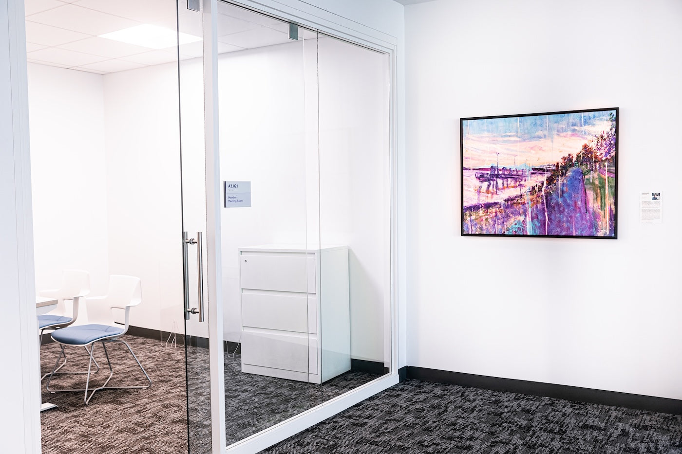  Displayed beside a glass-walled meeting room, Incantation by Ian Shearer depicts a shoreline at sunset in soft pinks, purples, and blues, with trees and water reflecting the fading light.