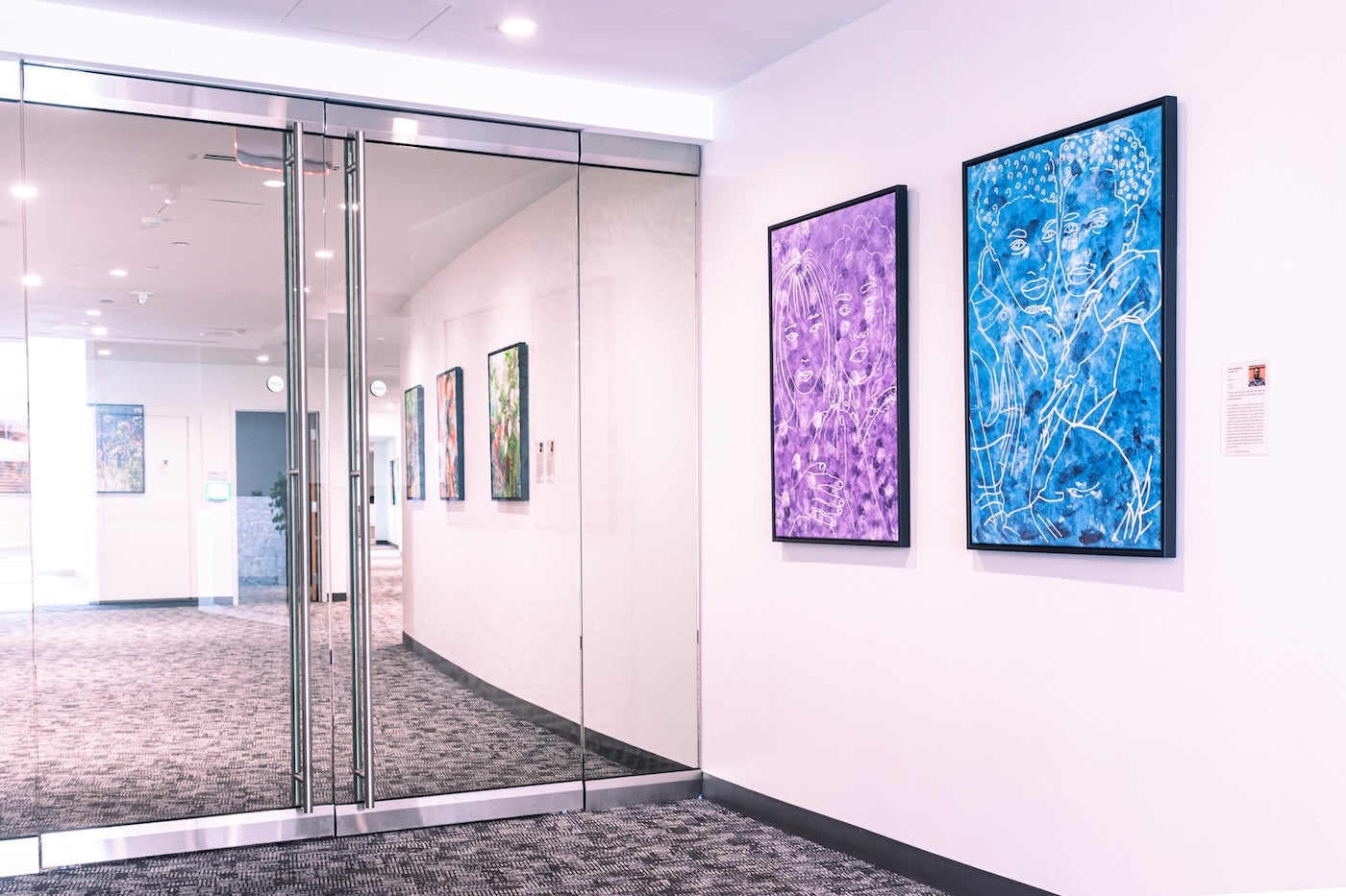Two figurative works by Lisa Murphy hang side by side. As One (left) shows a pair of faces outlined in white over a purple background, while Together (right) features embracing figures drawn in white on a deep blue surface.