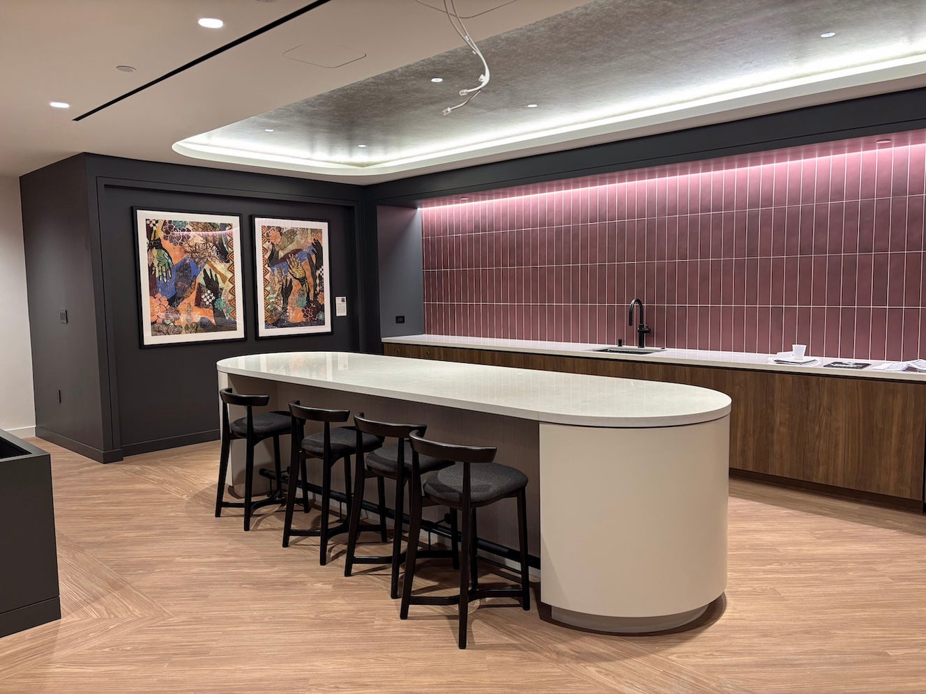 A modern lounge  at 777 6th St. NW features print reproductions of Karade’s Sankofa I and Karade’s Sankofa II by ArtLifting artist Diyah Najah, whose vibrant works add warmth and depth to the room’s contemporary design of curved white countertops, sleek dark cabinetry, and a mauve-tiled backsplash.