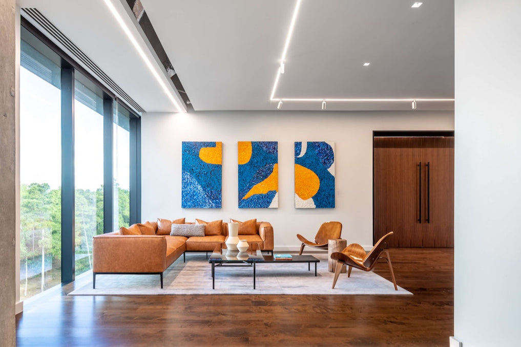 A contemporary executive office with leather seating and floor-to-ceiling windows. On the wall hangs a three-part series of original artworks by Marc, a triptych in bold blue and orange tones, placed prominently in the CEO’s office. 