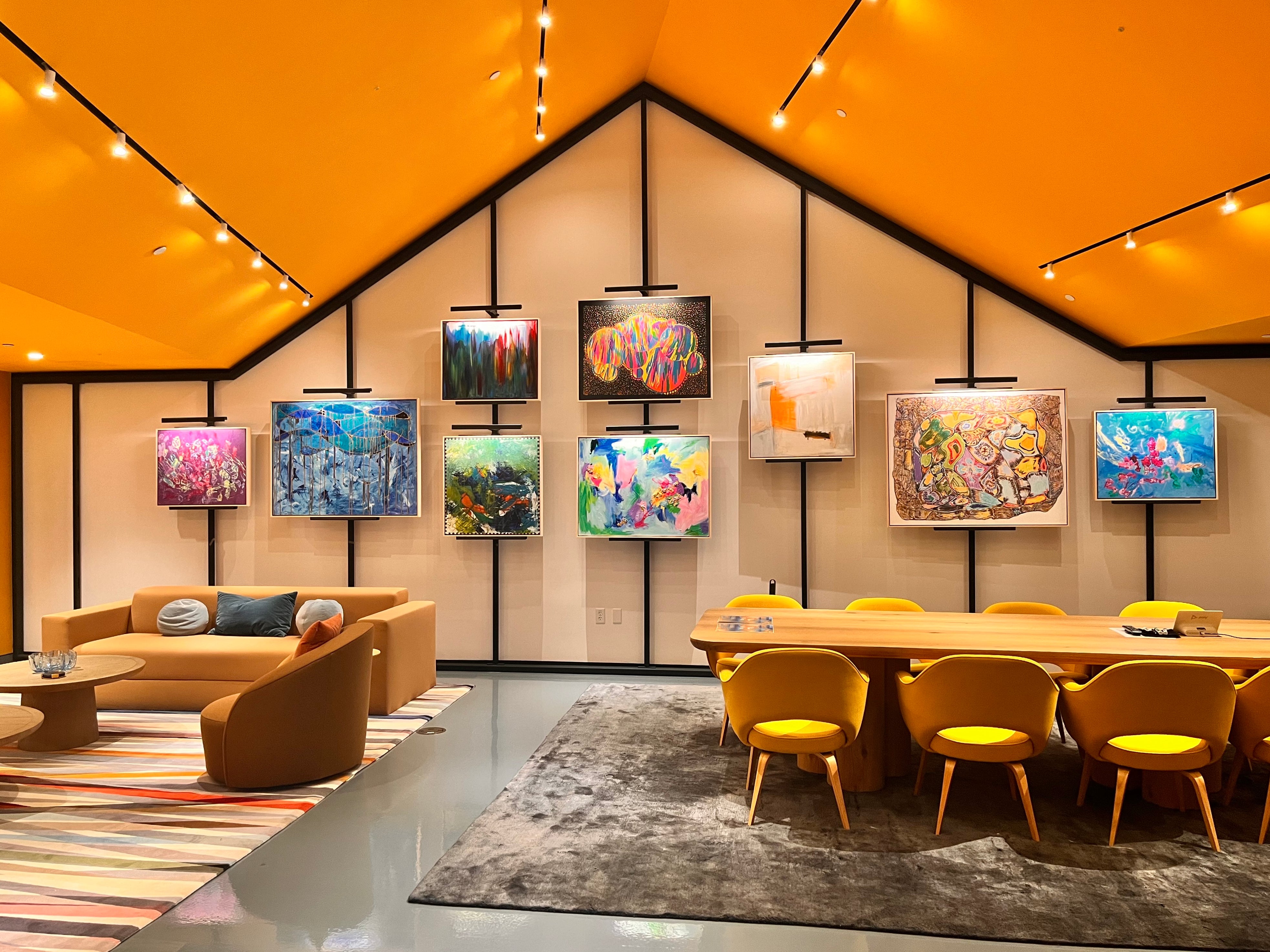 The image showcases "The Little House," a modern lounge featuring a unique gallery-style display of original artworks by ArtLifting artists. The space includes a long wooden table with yellow chairs, a tan seating area, and gallery catalogs on the table. Featured artists include: Mia Brown, Connie Avery, Quána Madison, Yvette, Sheila Smith, Aimee Hofmann, Charlie French, and Jeffrey Mayo. 
