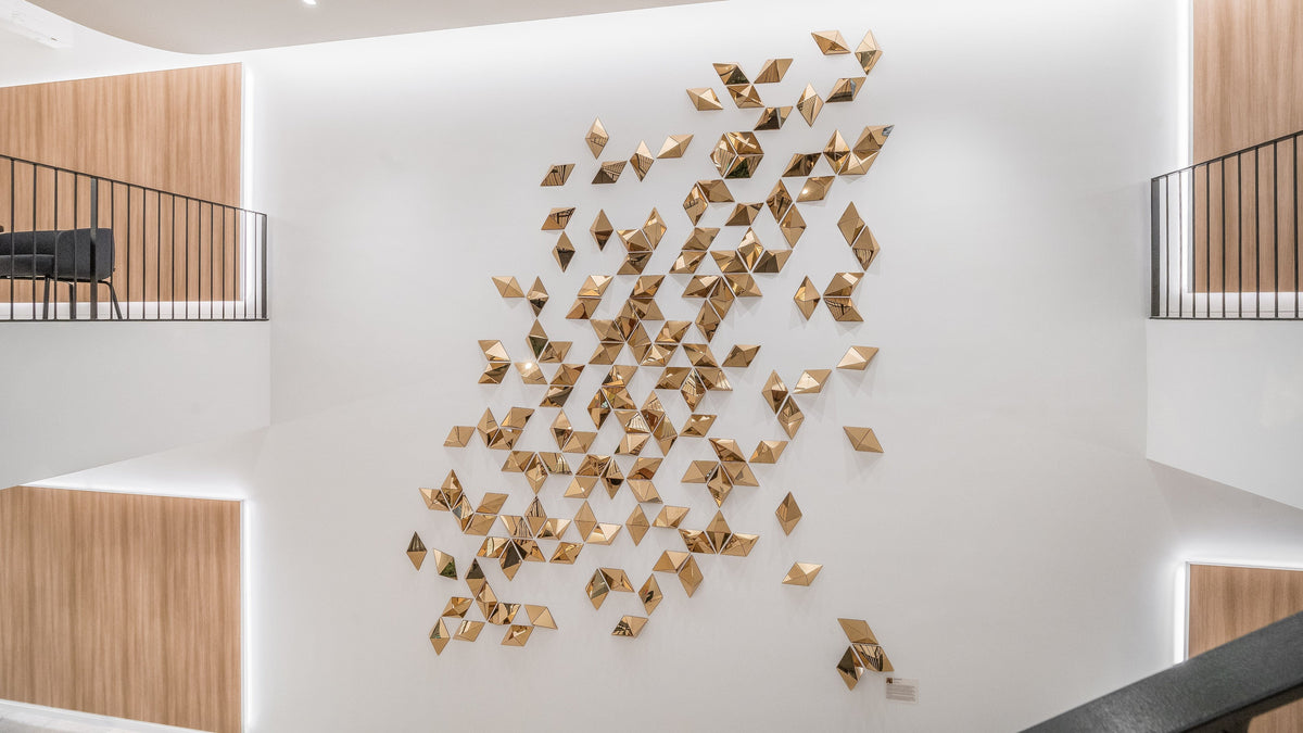 Eliza Redmann’s sculptural wall installation, Diamond Dreams, graces the lobby of JRM with its striking presence. Comprising 140 golden triangular pieces, the artwork cascades gracefully from near the ceiling to the base of a pristine white wall. Illuminated by overhead light fixtures, the installation radiates a captivating brilliance, creating a dynamic contrast against the lobby’s minimalist aesthetic. At the same time, its refined design harmonizes beautifully with the space's polished finishes.