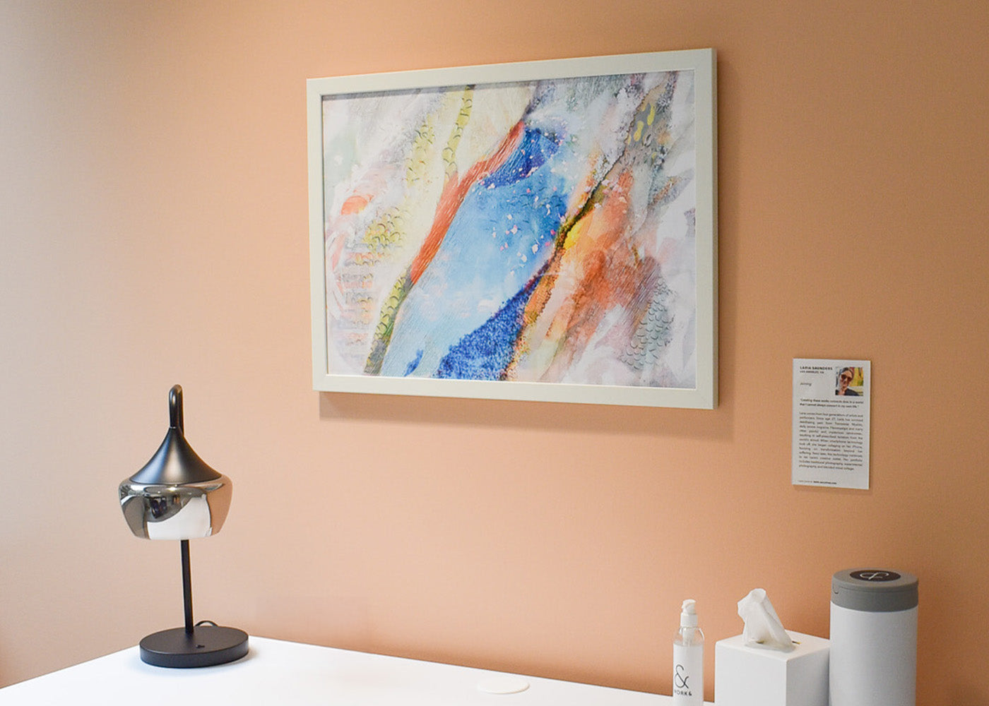 A framed fine paper print of Laria Saunders’s, Joining, is mounted on a peach-colored wall above a white desk in one of Work&’s wellness suites. 