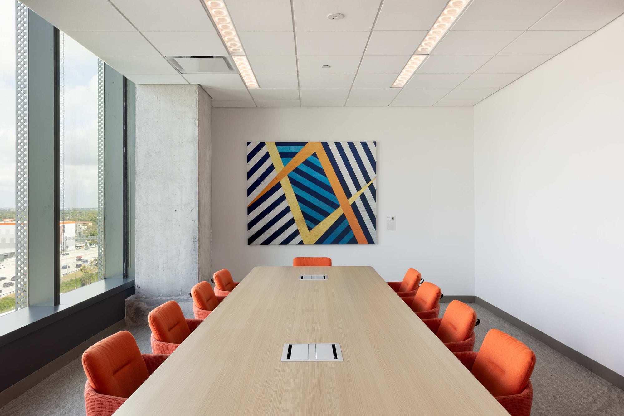 Modern conference room with a long table and orange chairs, featuring a geometric artwork on the wall.
