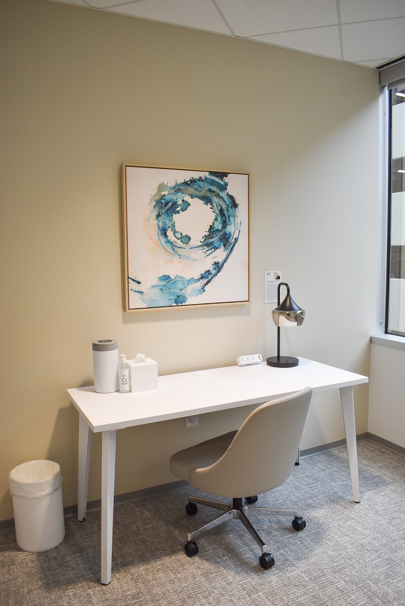 A framed canvas print of Christine Nelson’s, Wave, is mounted on a pale beige wall above a white desk in one of Work&’s wellness suites. 