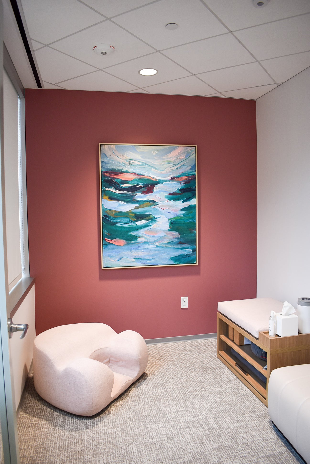 A framed canvas print of Elizabeth Shanahan’s, Trembling Waters, is mounted on a rose-colored wall. The space includes a lounge chair and a cushioned wooden bench with open storage arranged to foster quiet connection and restorative use in one of Work&’s wellness suites.