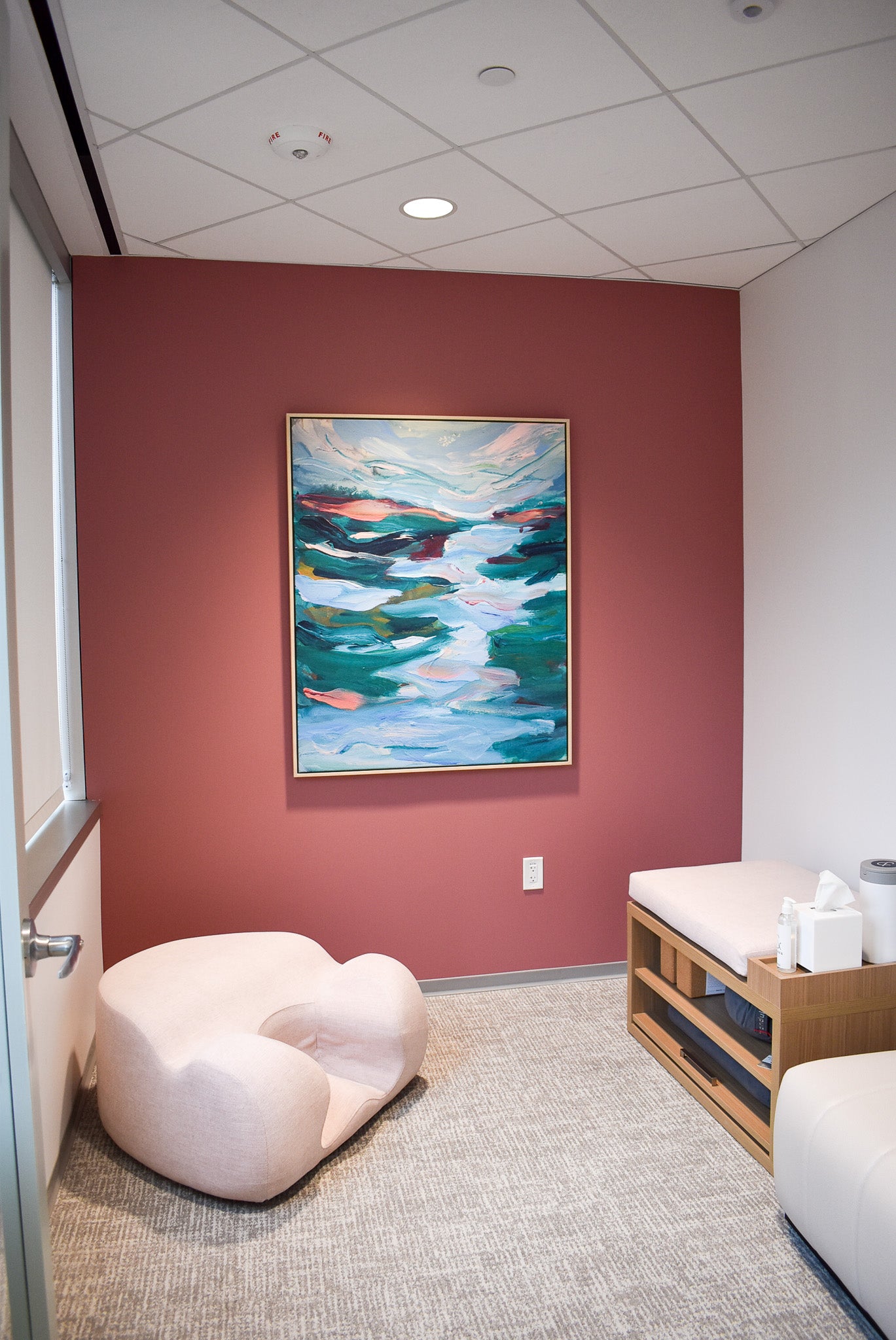 A framed canvas print of Elizabeth Shanahan’s, Trembling Waters, is mounted on a rose-colored wall. The space includes a lounge chair and a cushioned wooden bench with open storage arranged to foster quiet connection and restorative use in one of Work&’s wellness suites.