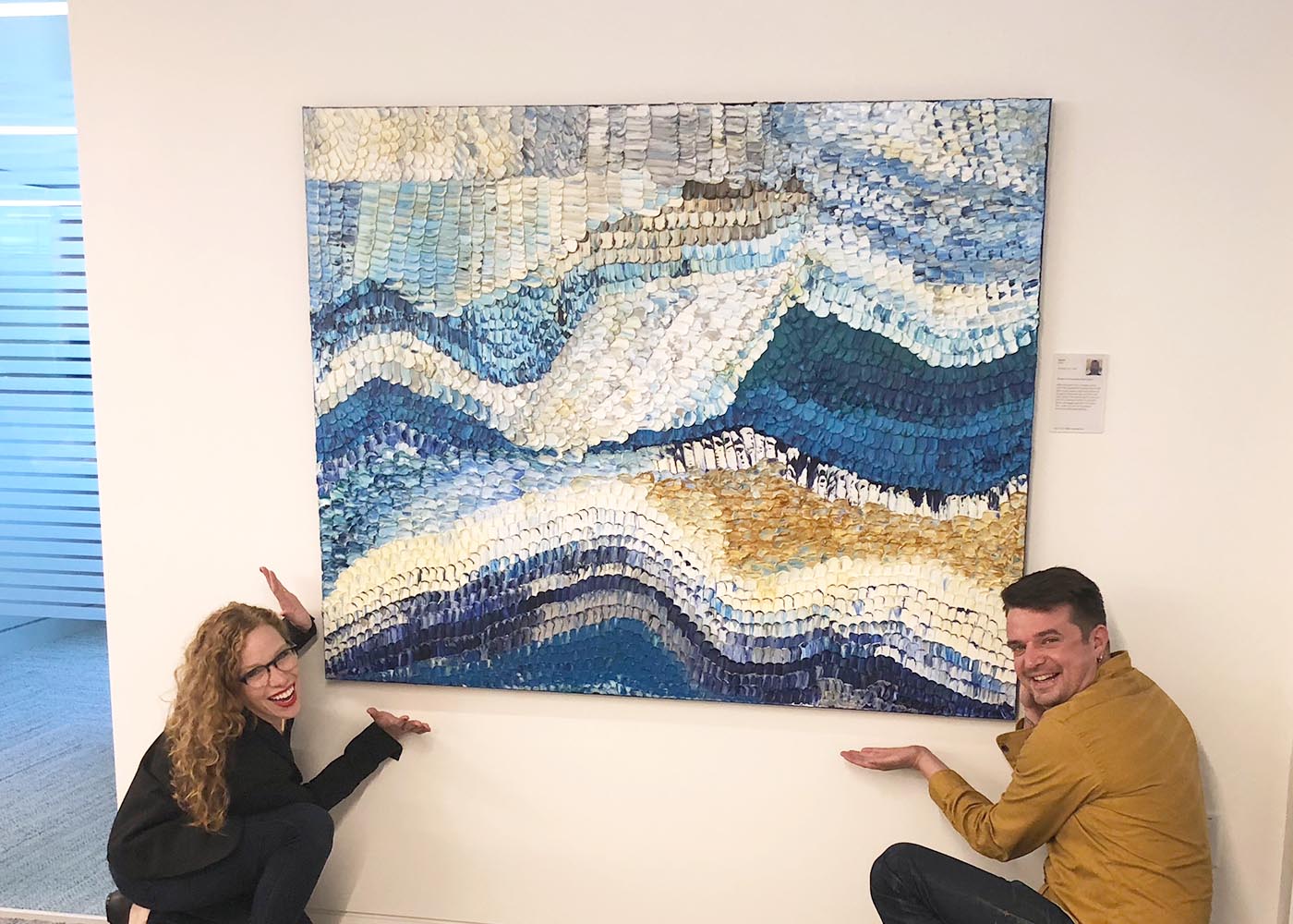 Two people posing in front of a large abstract artwork with wavy patterns in blue, white, and gold.